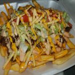 A Takeout Container Filled With Loaded Fries Topped With Ground Beef, Melted Cheese, Lettuce, Tomatoes, And Drizzled With A Creamy Sauce.
