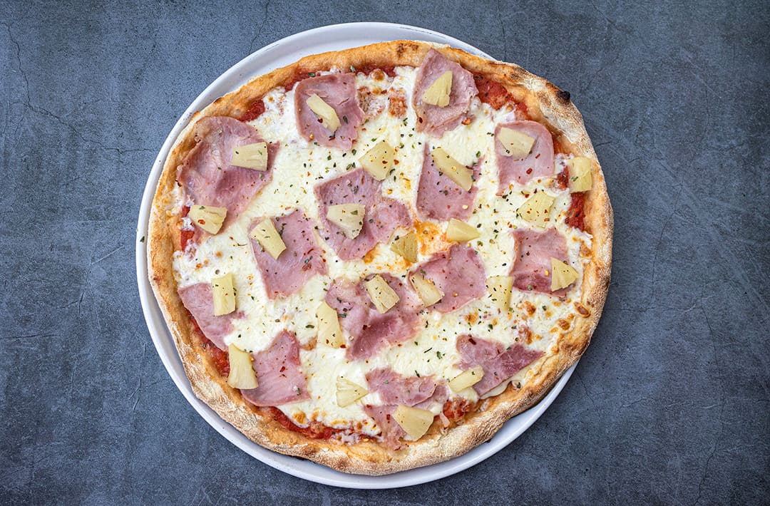 A freshly baked pizza topped with slices of ham, chunks of pineapple, and melted cheese on a white plate against a gray background.