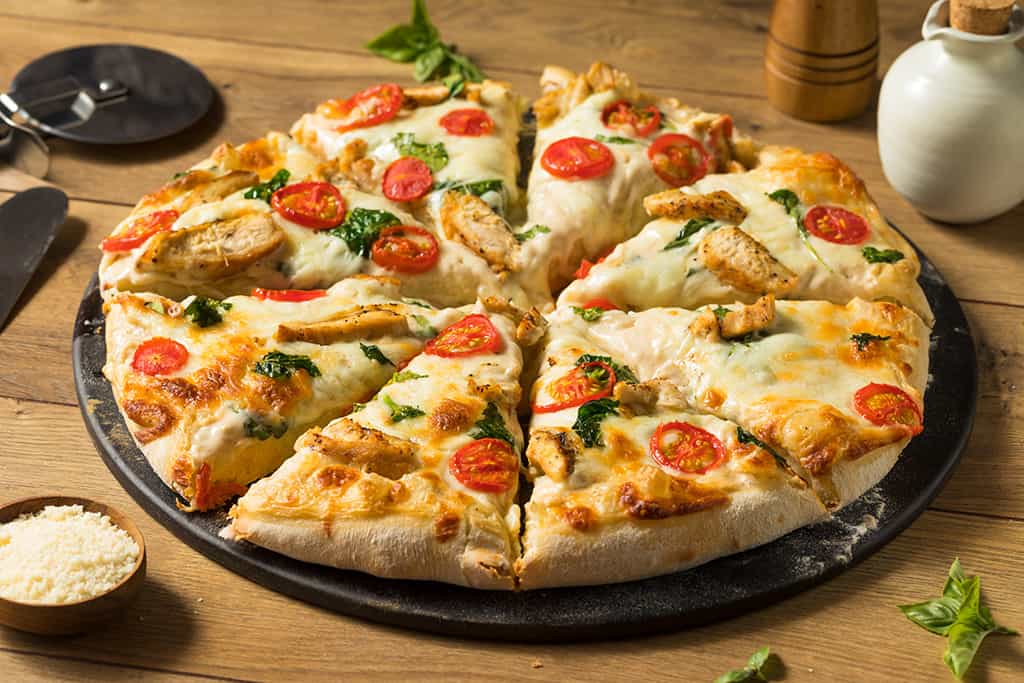 A sliced chicken pizza with tomatoes and spinach on a wooden table, accompanied by condiments.