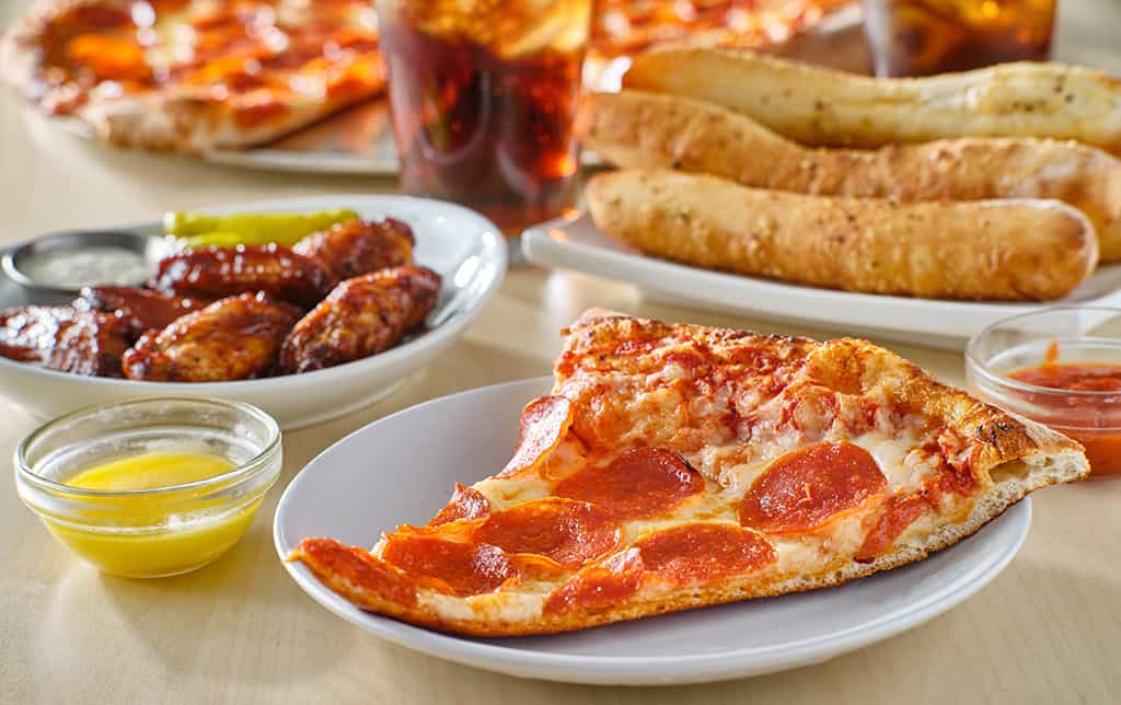 A table with a slice of pepperoni pizza on a plate, breadsticks, chicken wings, and dipping sauces, with more pizza and drinks in the background.