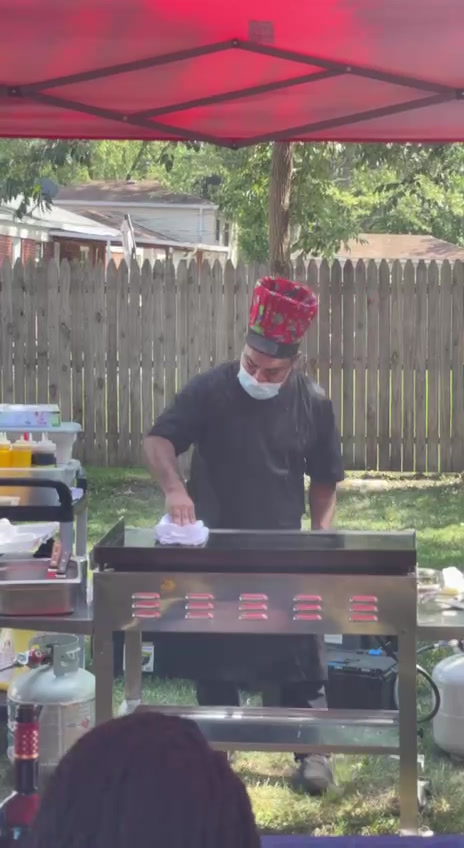 A Chef In A Festive Hat And Face Mask Cooking On A Grill Under A Red Canopy In A Backyard Setting.