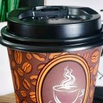 A disposable coffee cup with a steaming coffee illustration on a brown sleeve with coffee bean patterns, placed on a wooden surface with a plant and a partially visible sign in the background.