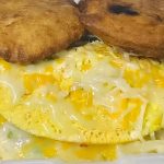 A foam container holding an omelet topped with melted cheese and mixed vegetables, served with two fried breads on top.