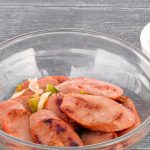 A glass bowl filled with slices of cooked sausage pieces and vegetables is placed next to a white cup containing a light-colored dipping sauce. The background is a dark textured surface.