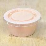 A small round container with a transparent lid, filled with a pinkish-beige substance, placed on a wooden surface.