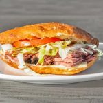 A sandwich on a plate, containing sliced meat, lettuce, tomato, and cheese inside a ciabatta roll, set on a wooden table.