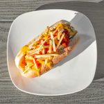 A hot dog topped with shredded cheese, ketchup, and crispy potato strips on a white plate on a gray wooden table.