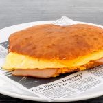 A sandwich with a thick, golden-brown bread, filled with an omelette and slices of ham, is placed on a white plate lined with newspaper-themed paper.
