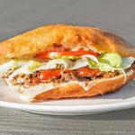 A sandwich with shredded meat, lettuce, tomatoes, and mayonnaise is served on a white plate.