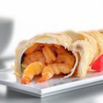 Close-up of a puff pastry filled with cooked shrimp, presented on a white rectangular plate. A red pepper garnish is visible beside it.