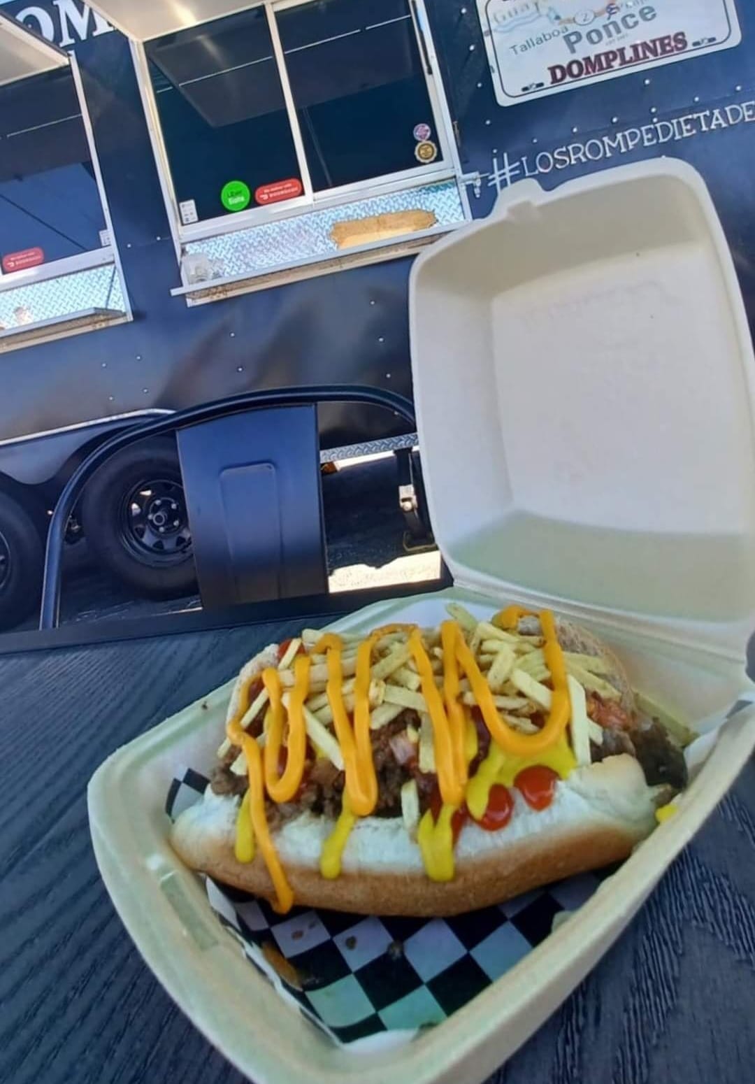 Close-up of a loaded hot dog with condiments in an open takeout container, placed on a wooden table outdoors, with a food truck in the background.