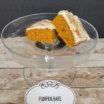 A Glass Cake Stand Displays Two Frosted Pumpkin Bars. A Label In Front Reads "pumpkin Bars" On A White Oval Card. The Background Is A Wooden Surface And A Dark Wall.