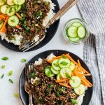 Two black plates with rice, seasoned beef, cucumber slices, and carrots, garnished with green onions and sesame seeds, next to a small bowl of cucumbers and a striped napkin.