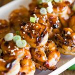 Grilled shrimp glazed with a sauce, garnished with sliced green onions and sesame seeds on a white plate.