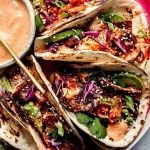 Close-up of three tacos filled with vegetables, slices of meat, sesame seeds, and garnished with fresh herbs. A bowl of dipping sauce is partially visible on the side.