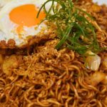 A plate of fried noodles topped with a sunny-side-up egg, garnished with sliced green onions and fried shallots.