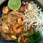 A bowl of shrimp pad Thai garnished with chopped peanuts, bean sprouts, green onions, chili flakes, and a lime wedge.