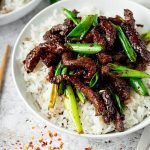 A bowl of rice topped with strips of cooked beef and vibrant green onions.