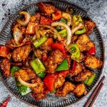 A plate of stir-fried chicken with bell peppers and onions, garnished with spices. Chopsticks are placed beside the plate.