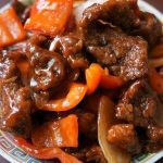 A plate of cooked beef strips with red bell peppers and onions in a savory sauce.