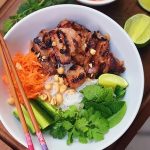 Bowl of Vietnamese noodle salad with grilled pork, rice noodles, carrots, cucumber, mint, coriander, peanuts, lime wedges, and chopsticks on the side.