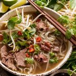 A bowl of beef pho with rice noodles, bean sprouts, herbs, chili slices, and chopsticks.