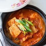 A bowl of kimchi jjigae with tofu, green onions, and various vegetables, served in a black pot with a partially open decorated lid.