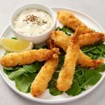 A plate of breaded shrimp on a bed of greens with a lemon wedge and a small bowl of dipping sauce on the side.
