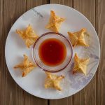 Plate of six fried wontons arranged in a circle around a bowl of red dipping sauce on a wooden table.