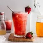 A tall glass of strawberry smoothie with a metal straw and two whole strawberries, placed on a wooden board. In the background, there are glass bottles and jars.