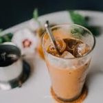 Glass of iced coffee with a metal straw on a coaster, surrounded by blurred greenery and flowers in the background.