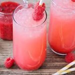 Two glasses of pink raspberry lemonade with ice and straws, garnished with fresh raspberries. A jar of raspberry puree and loose raspberries are on a wooden surface nearby.