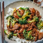 A dish of beef and broccoli with brown sauce served over a bed of white rice, accompanied by wooden chopsticks on the side.