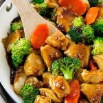 A skillet with stir-fried chicken, broccoli, and carrots in a glossy sauce, with a wooden spatula.