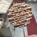 Hand Holding A Chocolate Covered Lollipop With White Drizzle And Chopped Nuts, In A Kitchen Setting With Trays In The Background.