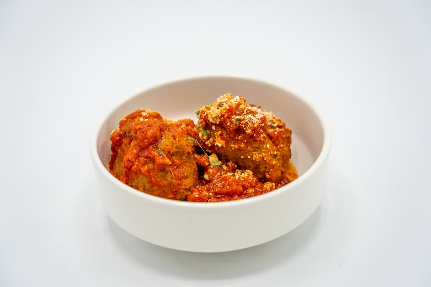 Two meatballs with marinara sauce and a sprinkling of parmesan cheese in a white bowl against a plain white background.