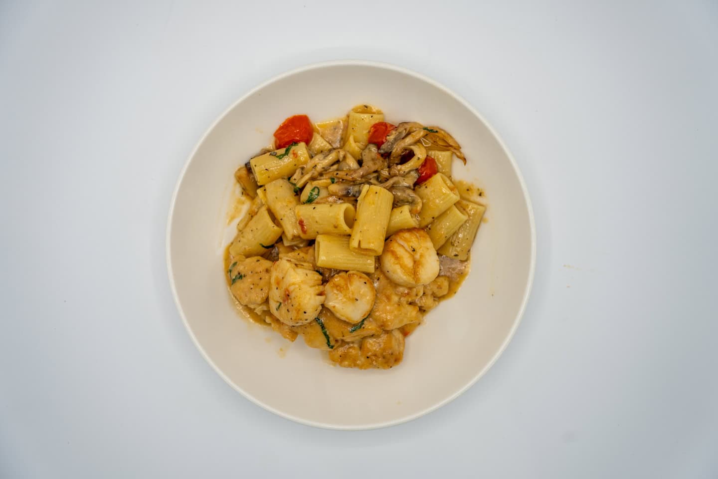 A plate of rigatoni pasta with shrimp, mushrooms, zucchini, and red peppers, served in a light sauce on a white background.