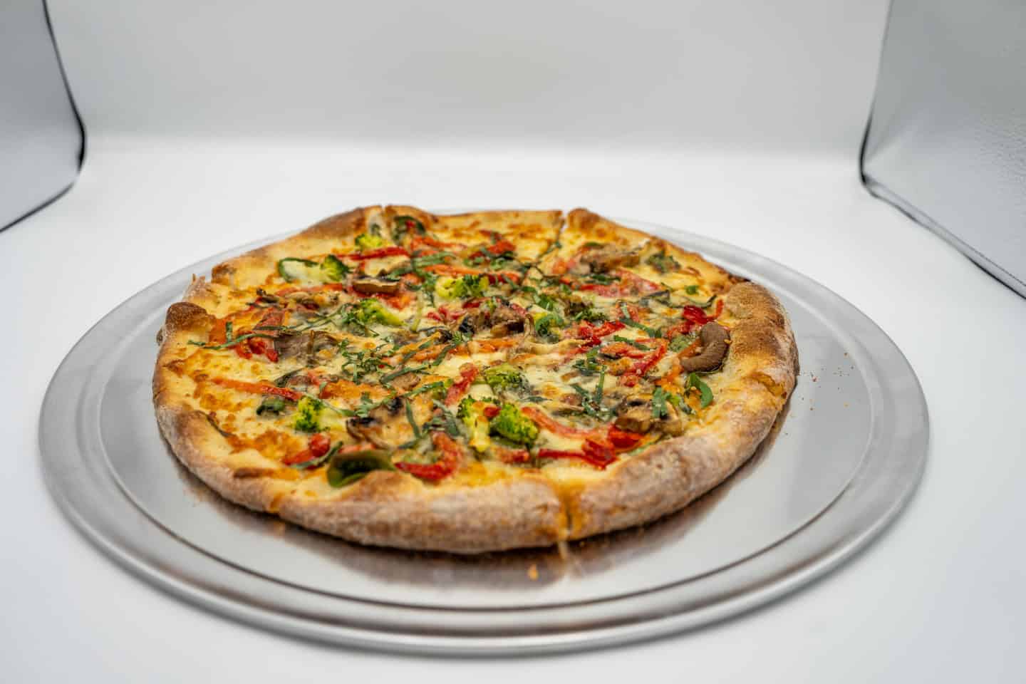 A whole, freshly baked pizza with various toppings on a metal plate, displayed in a lightbox setting.