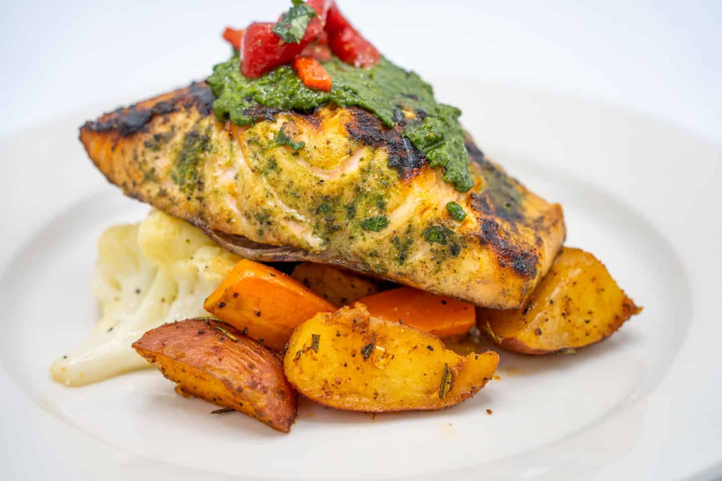 Grilled salmon fillet topped with green herb sauce, served with roasted carrots and a side of mashed potatoes on a white plate.