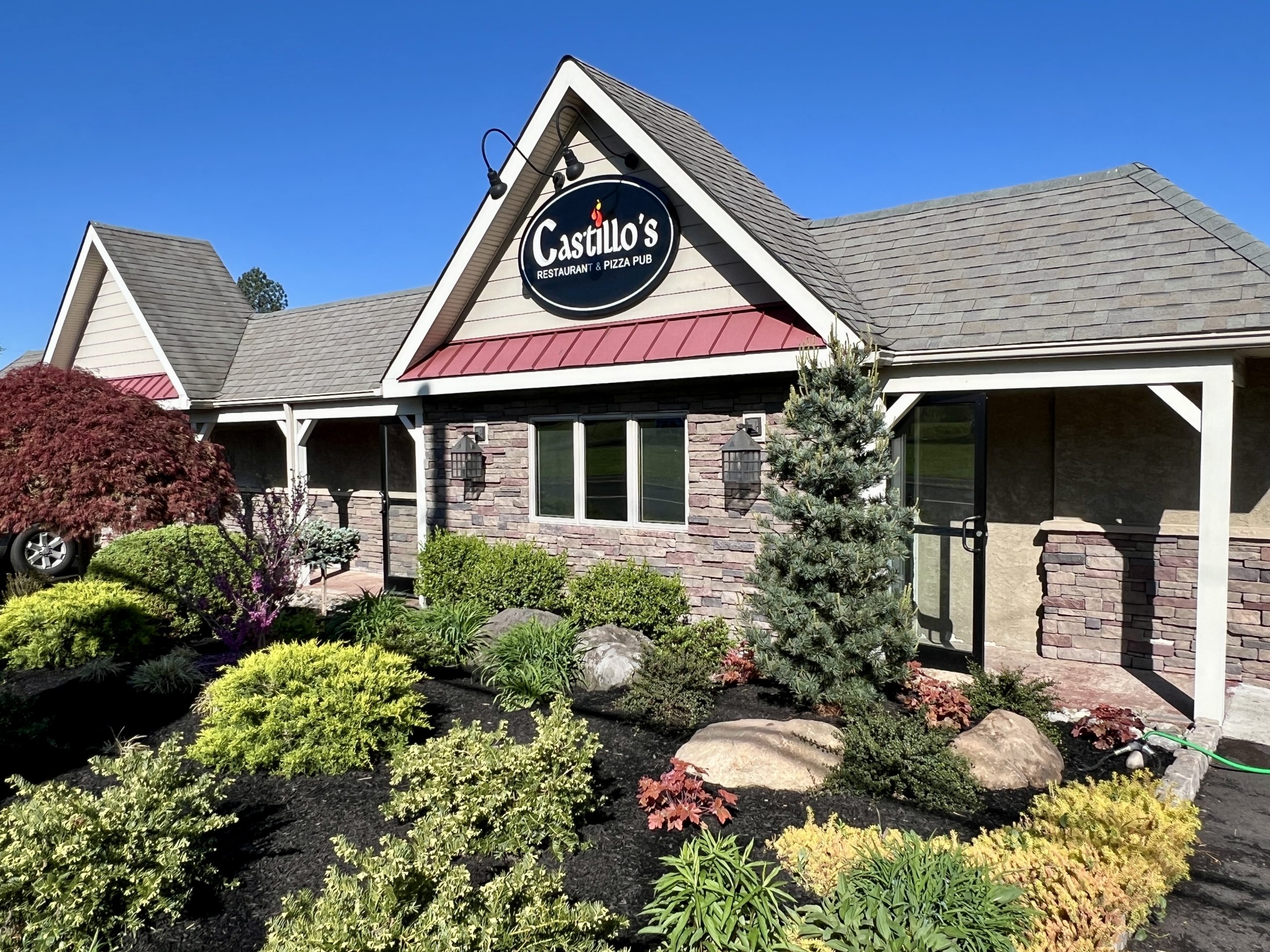 Exterior of castillo's mexican restaurant featuring stone and siding facade, landscaped with shrubs and small trees under a clear blue sky.