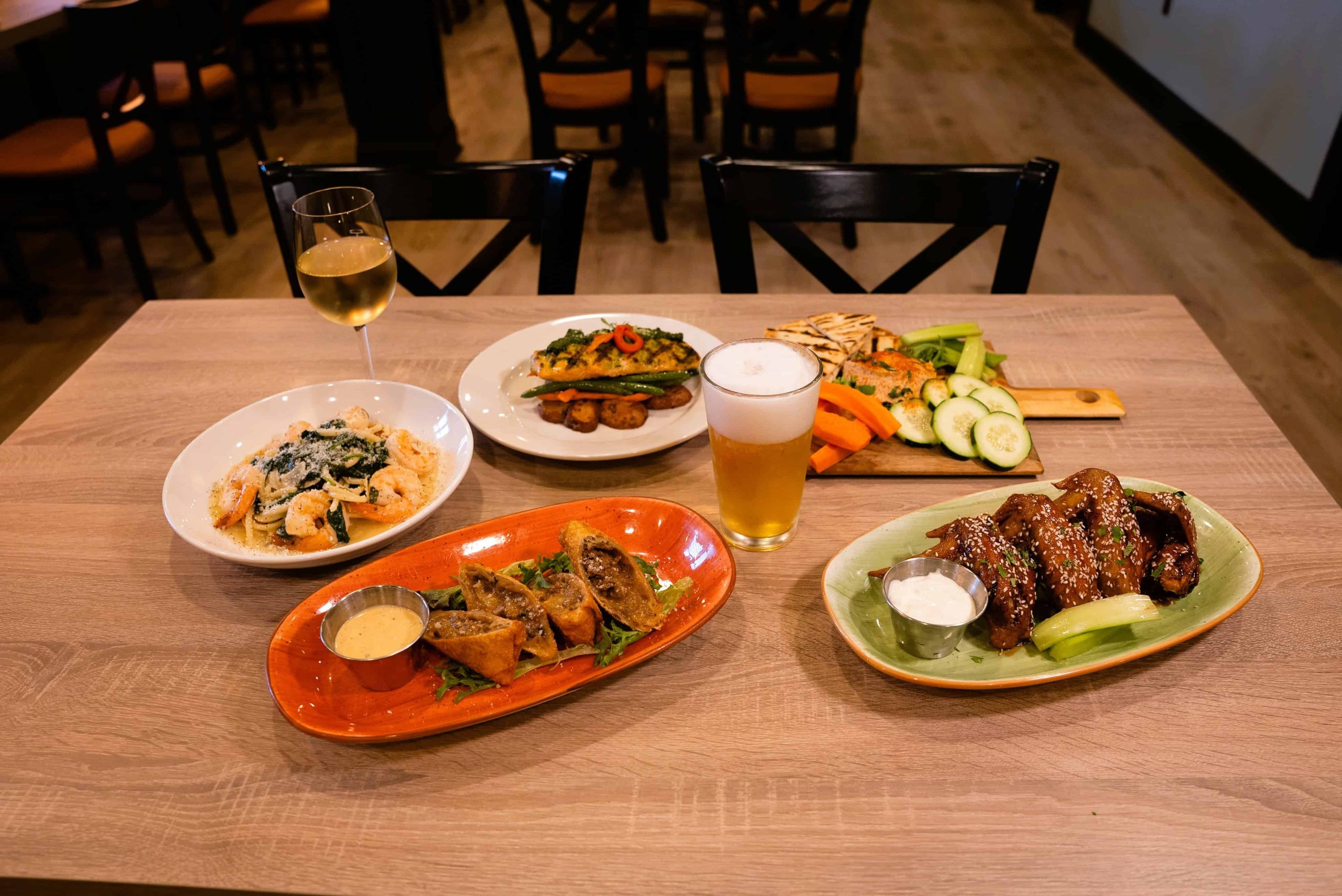 A table with various dishes including pasta, spring rolls, chicken wings, grilled vegetables with bread, a glass of white wine, and a pint of beer.