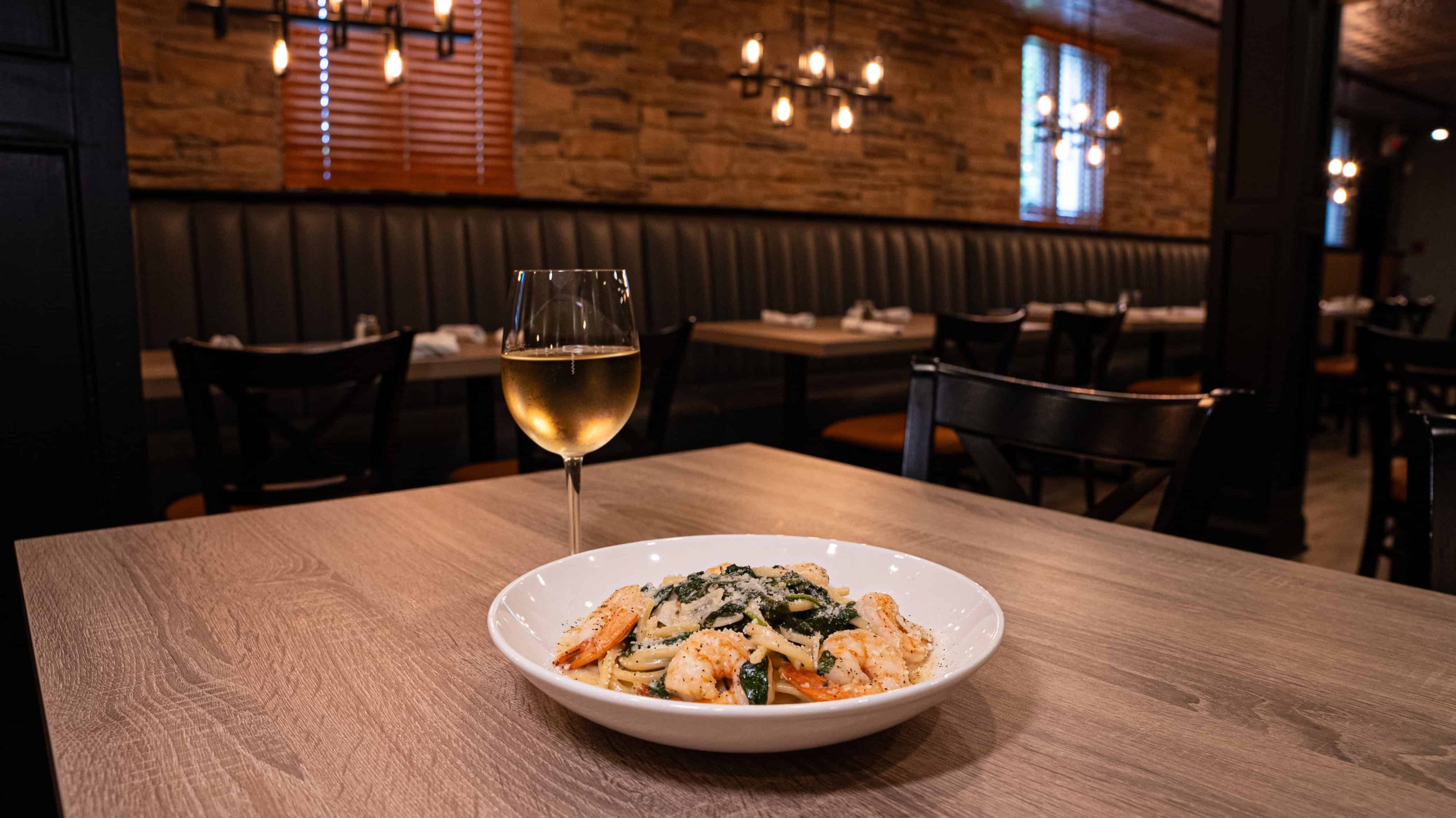 A plate of pasta with shrimp and vegetables served on a wooden table with a glass of white wine in a dimly lit restaurant.