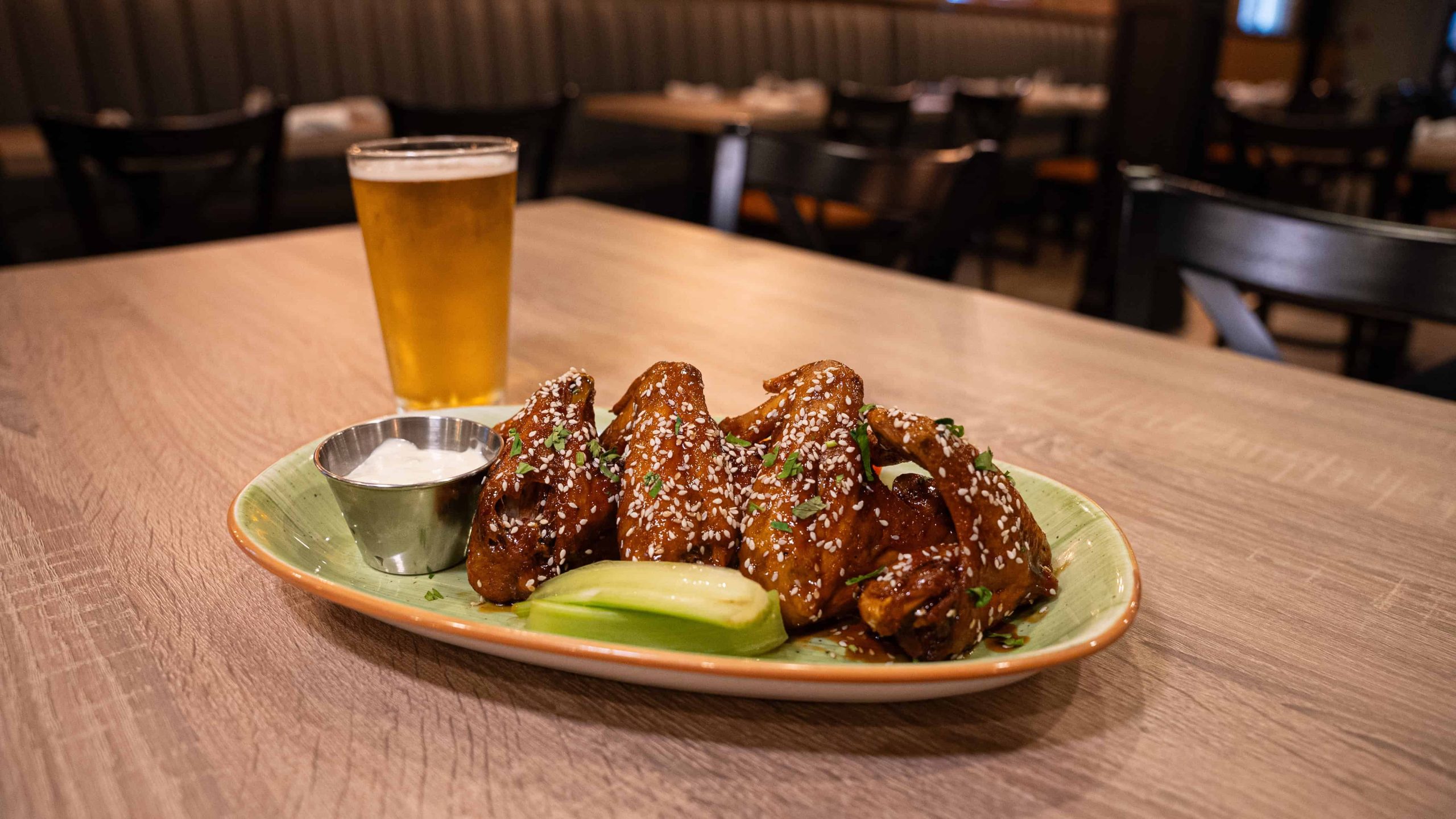 Plate of sesame-covered chicken wings with celery, ranch sauce, and a glass of beer on a wooden table in a restaurant. Empty chairs and tables are visible in the background.