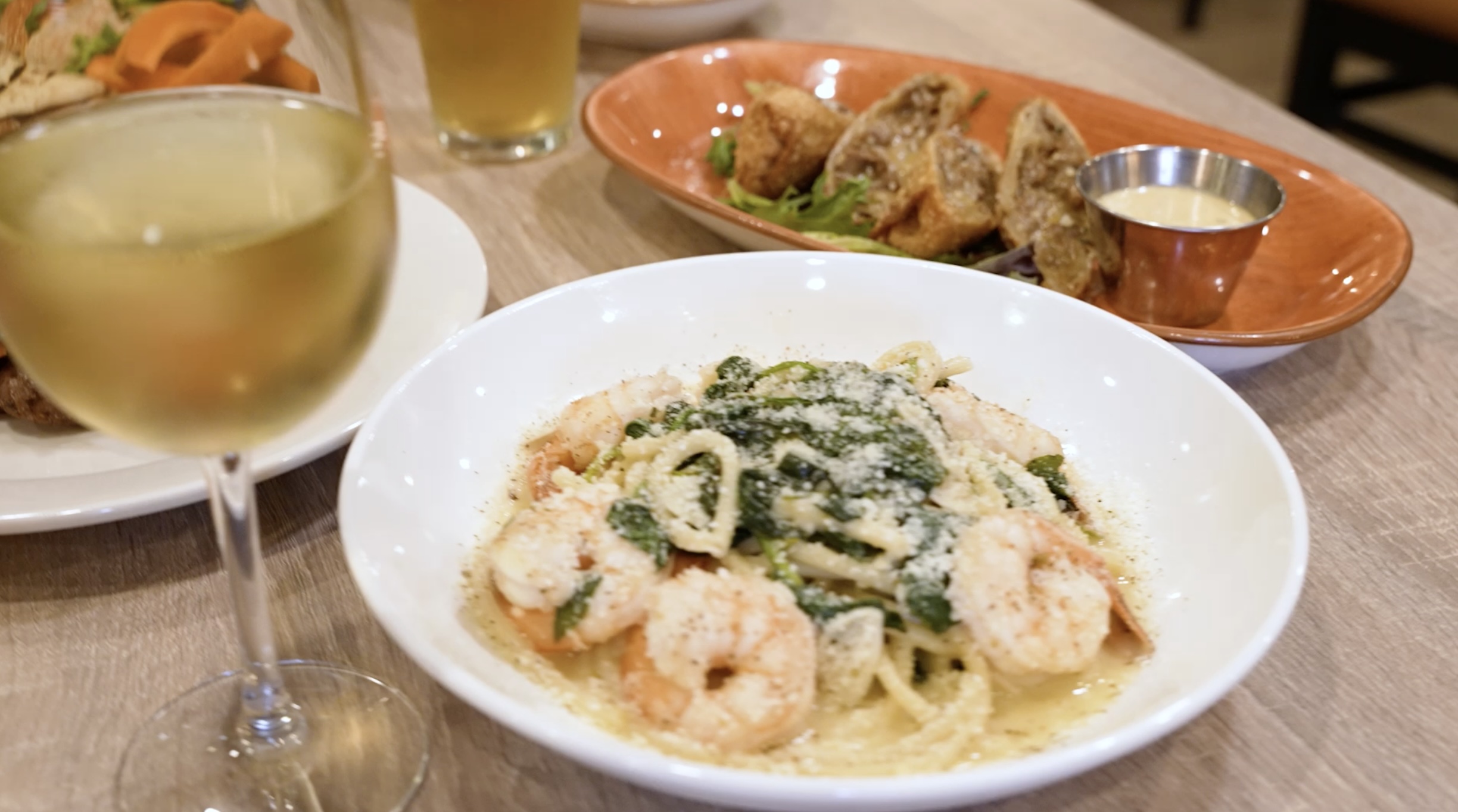 A bowl of shrimp pasta with greens topped with grated cheese, flanked by a glass of white wine and a plate of appetizers on a wooden table.