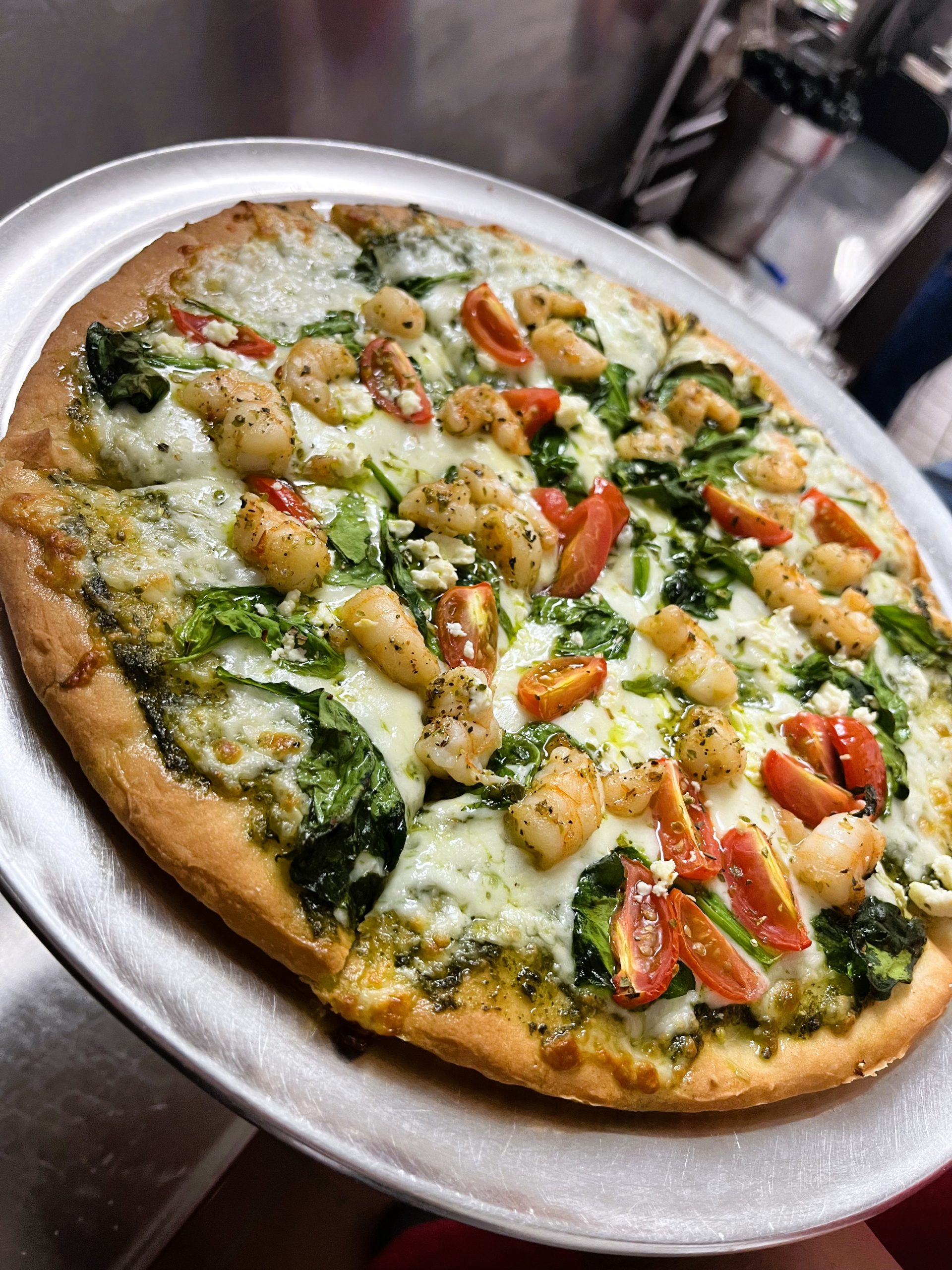 A fresh pizza on a metal tray topped with shrimp, spinach, cherry tomatoes, and melted cheese.