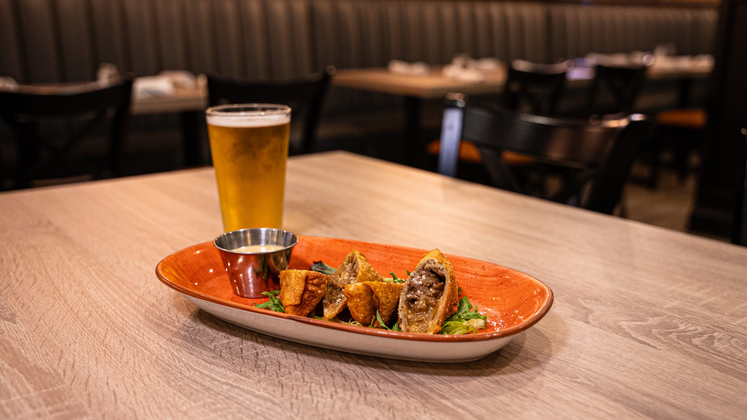 A glass of beer and a plate of food with fried rolls and dipping sauce on a wooden table in a restaurant setting.