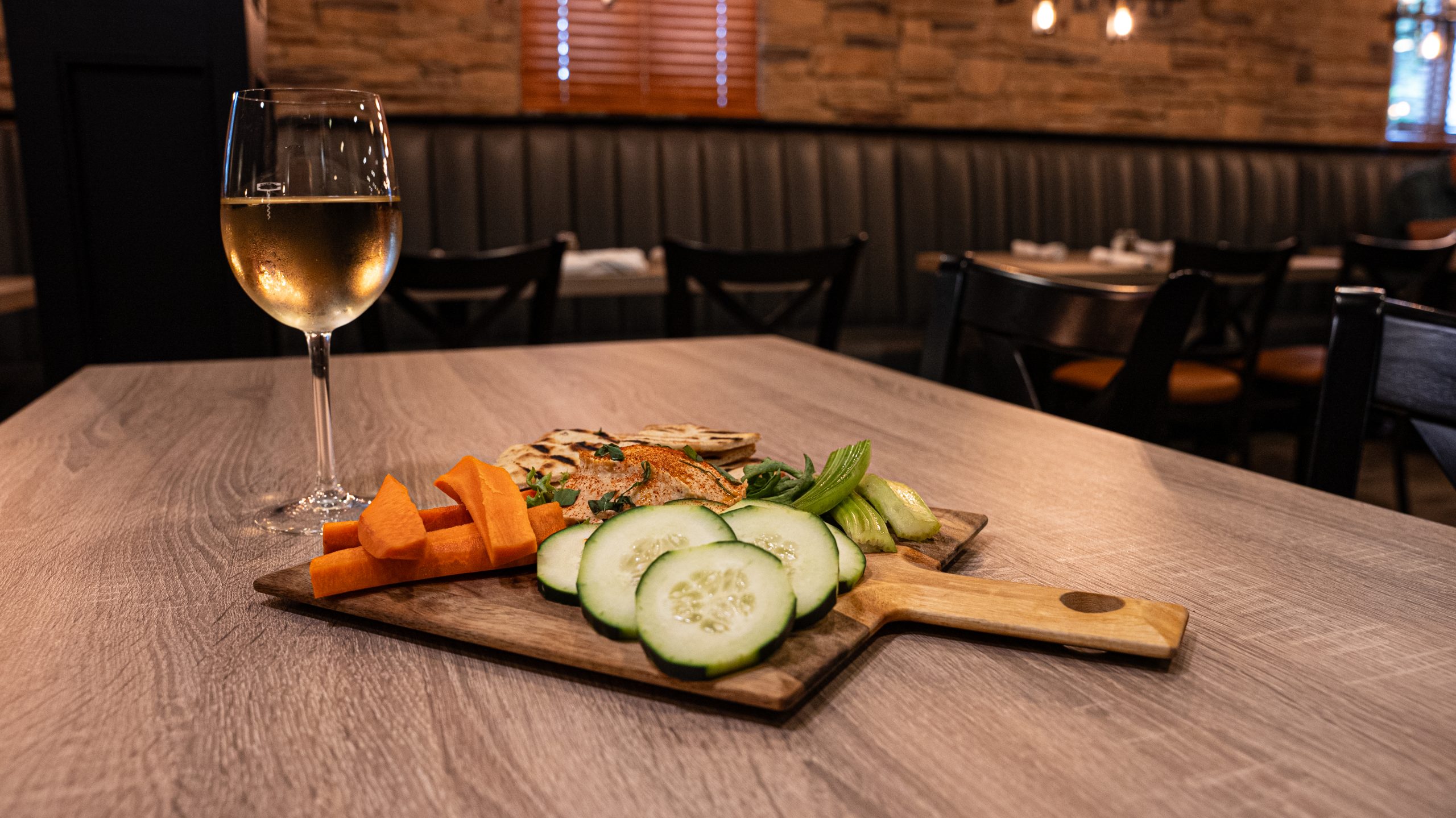 A wooden board with sliced cucumbers, carrots, and pita bread sits on a table alongside a glass of white wine in a restaurant setting.