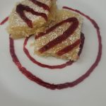 Two Square Pastries Dusted With Powdered Sugar, Drizzled With Red Sauce, And Placed On A White Plate With A Circular Sauce Pattern.