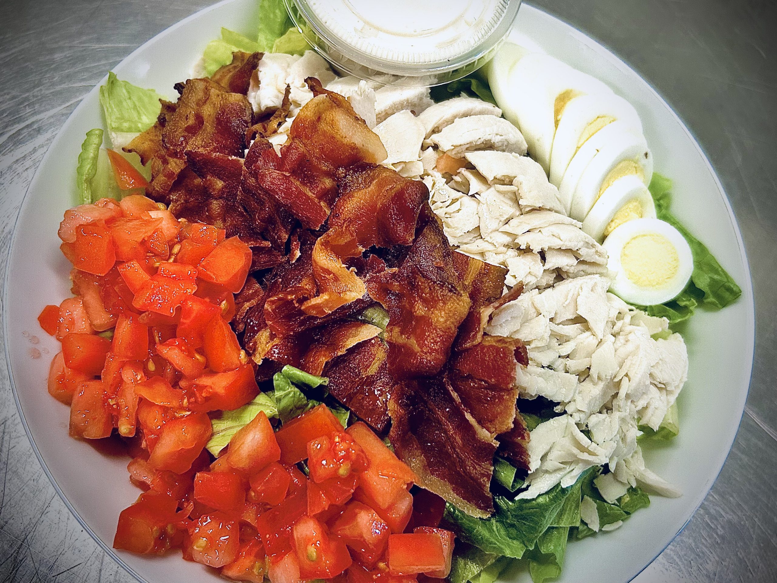 A salad bowl with chopped lettuce, diced tomatoes, bacon strips, sliced chicken breast, hard-boiled egg slices, and a container of dressing on the side.