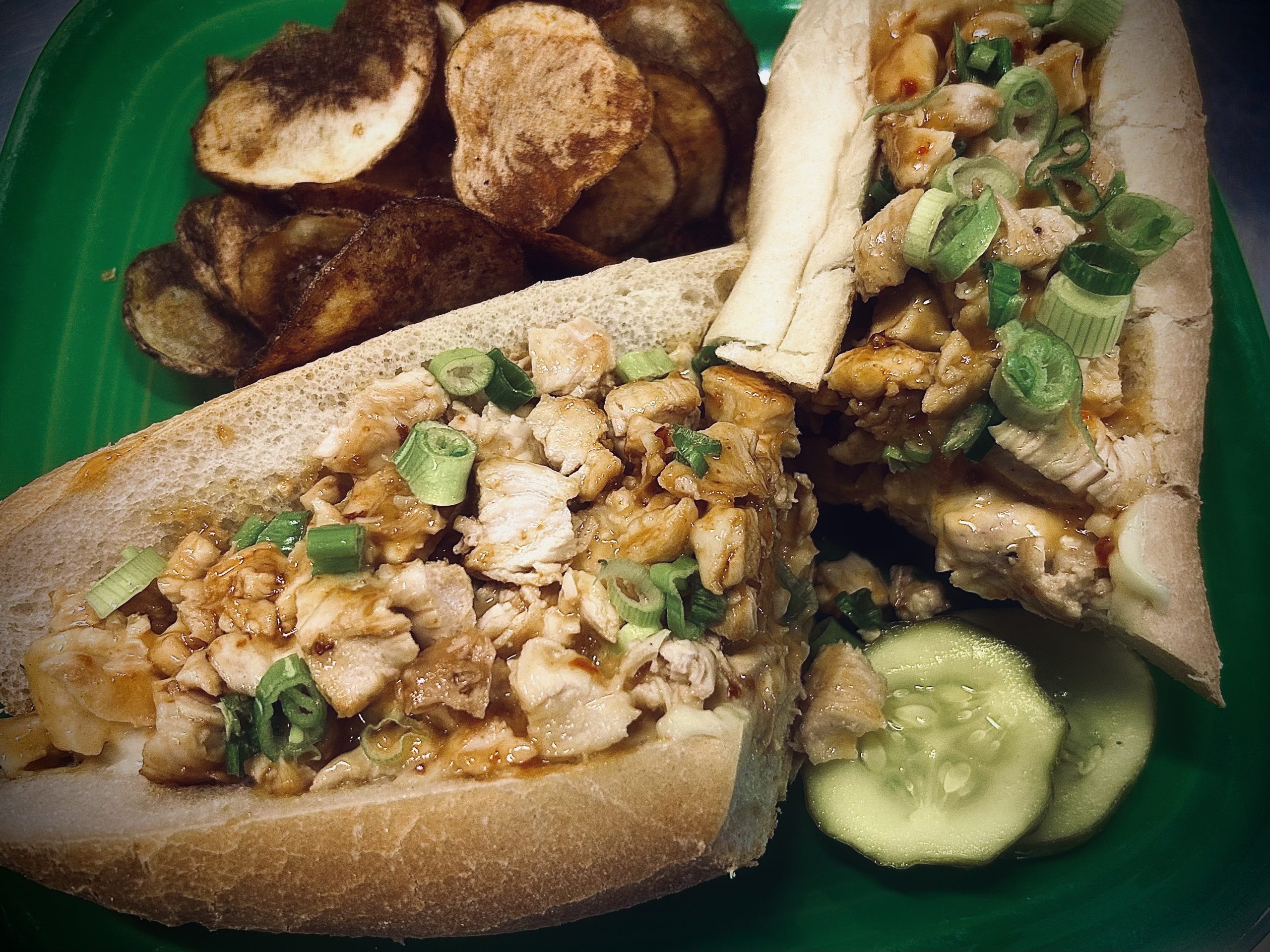 Two sandwiches filled with chopped chicken and green onions, served with sliced pickles and a side of potato chips on a green plate.